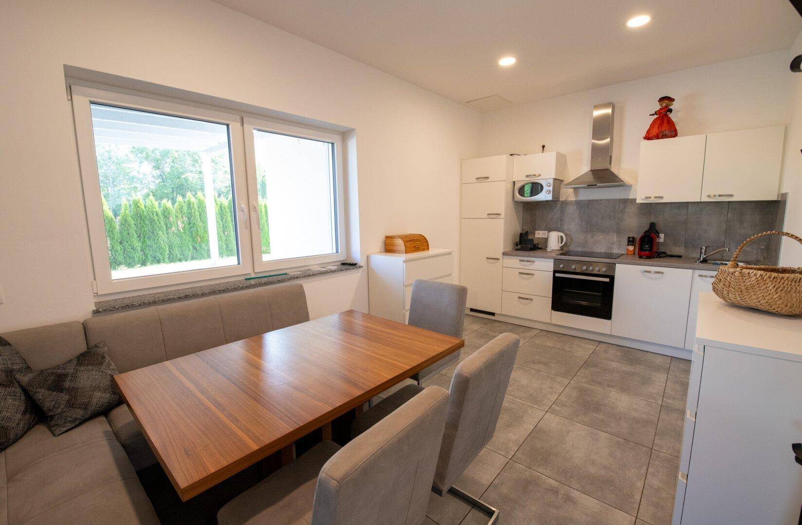 Moderne Wohnküche mit weißer Einbauküche, Esstisch und Blick ins Grüne durch das Fenster.