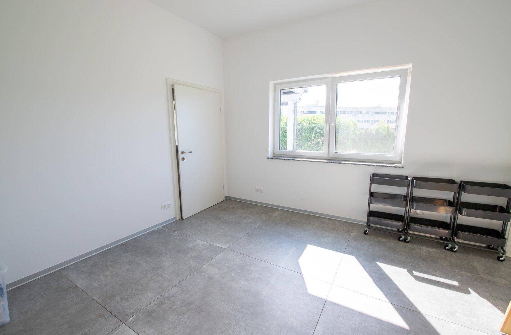 Leerer, heller Raum mit grauen Fliesen und einem Fenster, ideal für verschiedene Nutzungen.