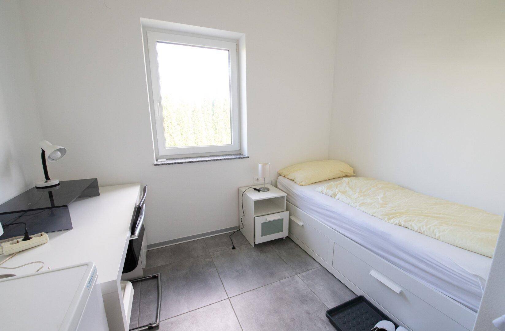 Kleines Schlafzimmer mit Einzelbett, Schreibtisch und Fenster, hell und funktional eingerichtet.