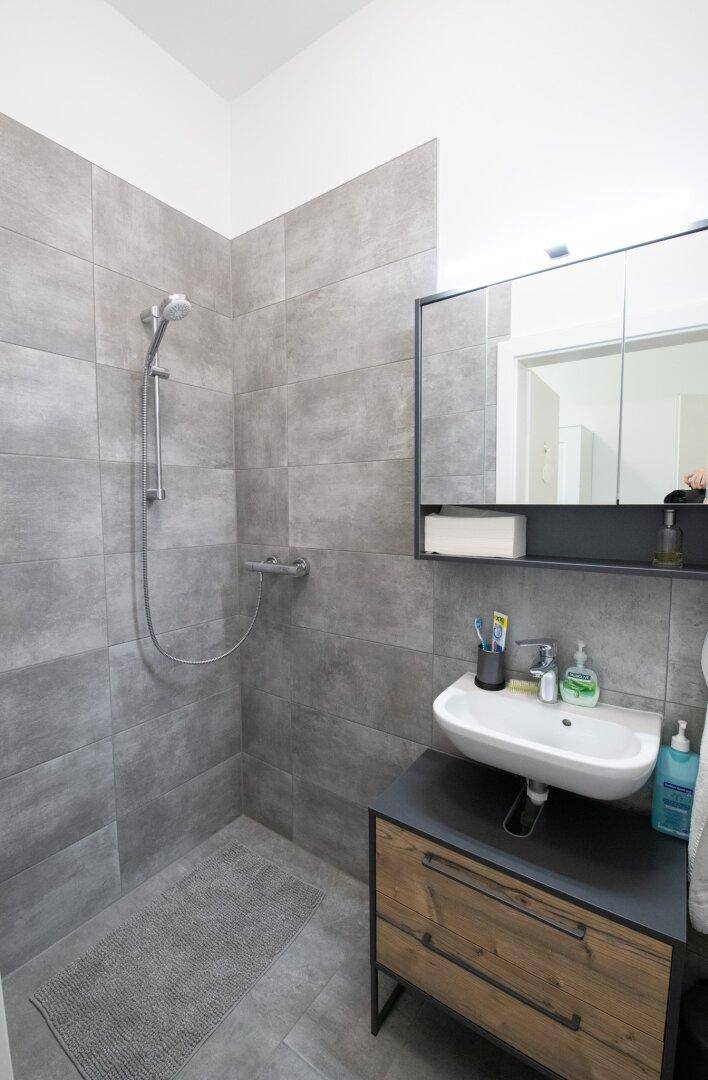 Modernes Badezimmer mit grauen Großformatfliesen, ebenerdiger Dusche und Waschtischunterschrank.