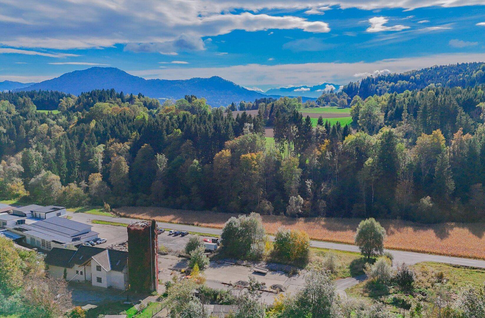 Weitläufige Landschaftsaufnahme mit Bergen, Wäldern und einem kleinen Industriegebiet im Vordergrund unter blauem Himmel.