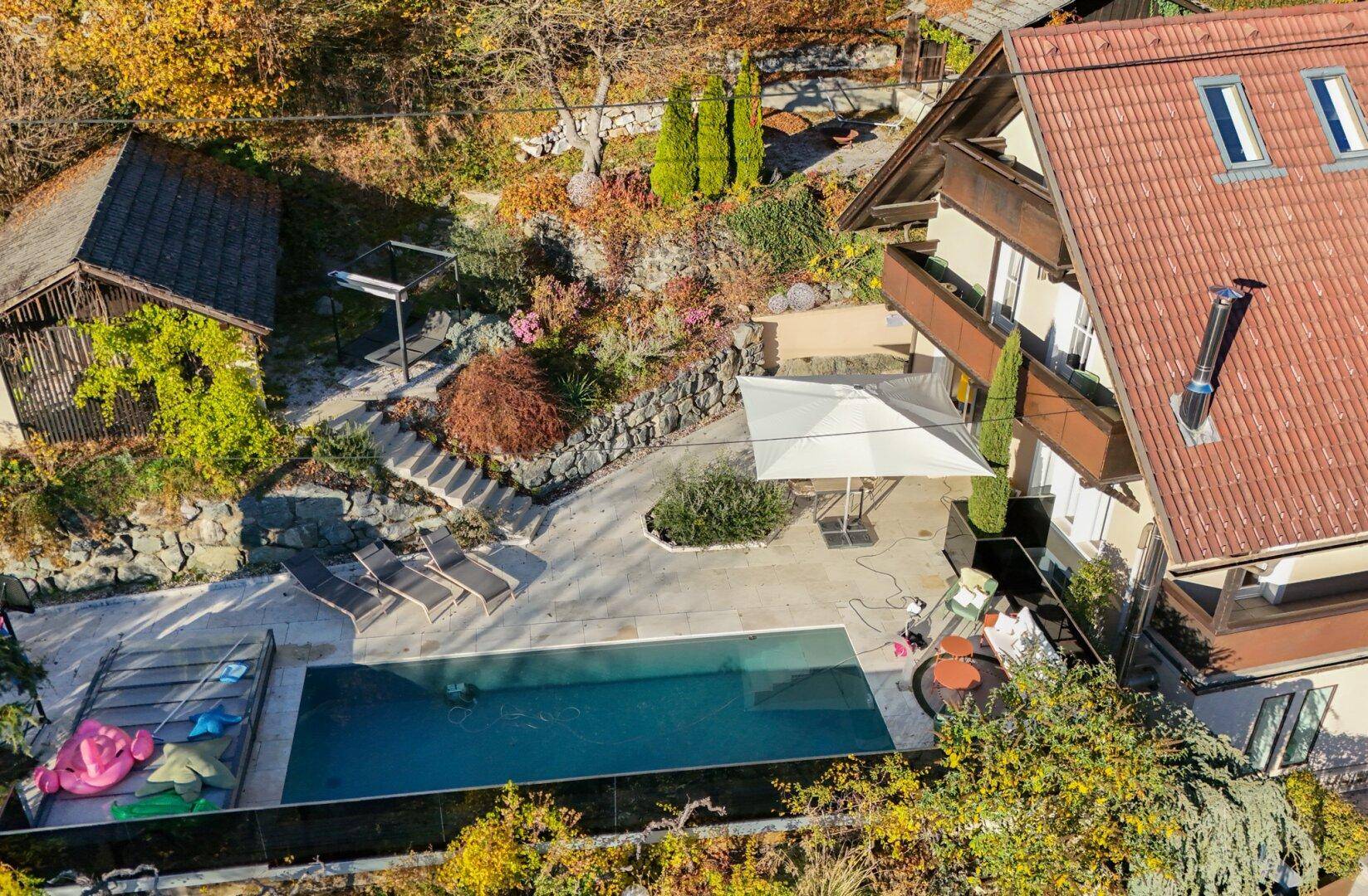 Detailansicht des Außenbereichs mit Pool, Liegestühlen und Sonnenschirm, umgeben von Natur.