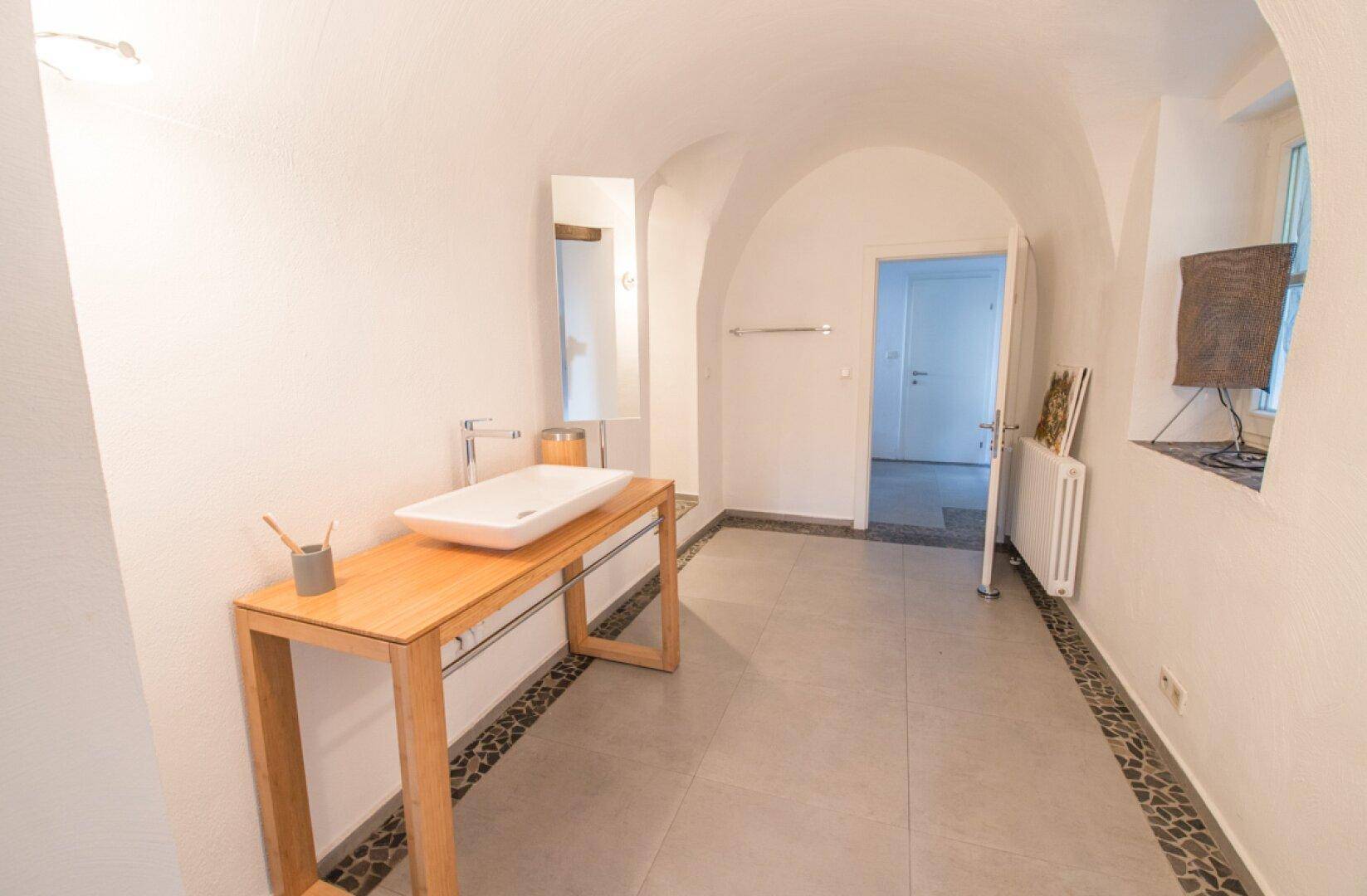 Heller Badezimmerbereich mit modernem Waschbecken, Spiegel und gewölbten Decken, führt zu einem weiteren Raum.