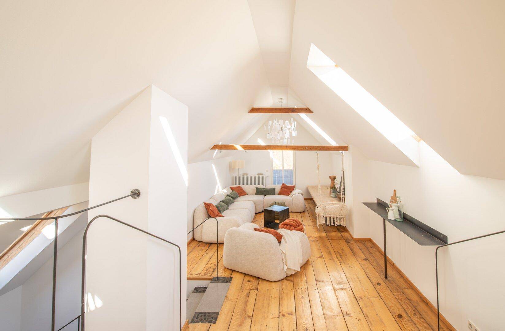 Geräumiges Dachgeschoss-Wohnzimmer mit schrägen Decken, Holzbalken und Blick auf die Treppe.