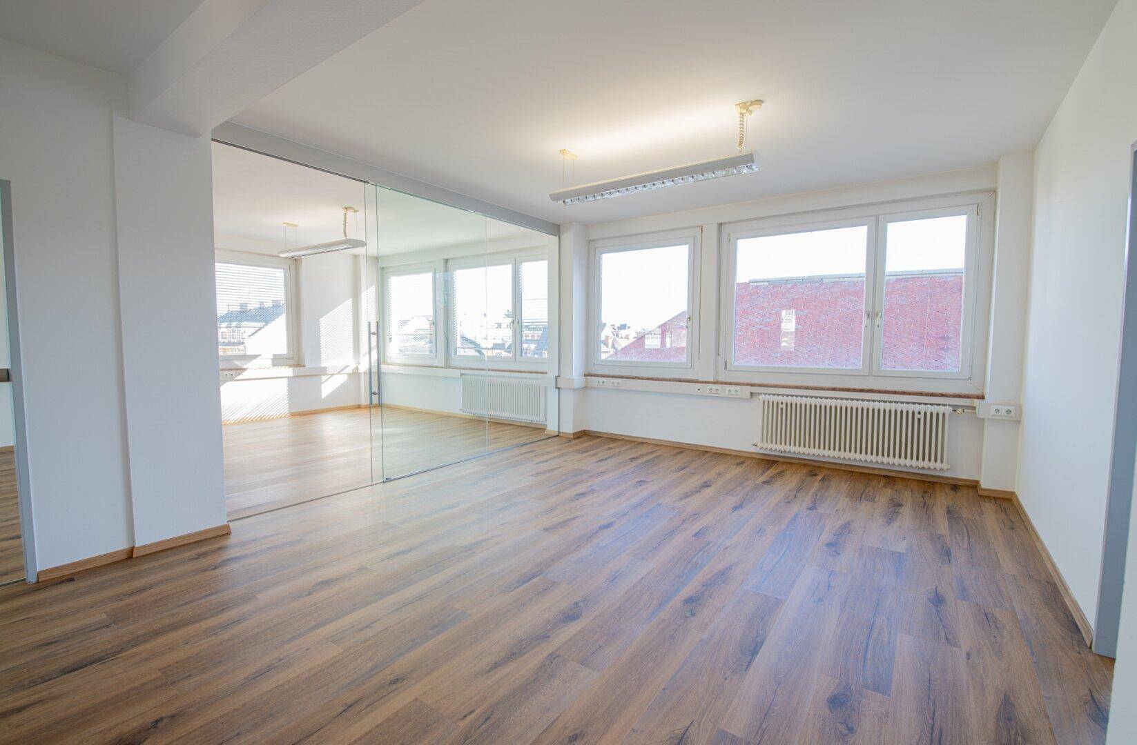 Heller Büroraum mit großen Fenstern, Laminatboden und einer Glaswand, die einen weiteren Bereich abtrennt.