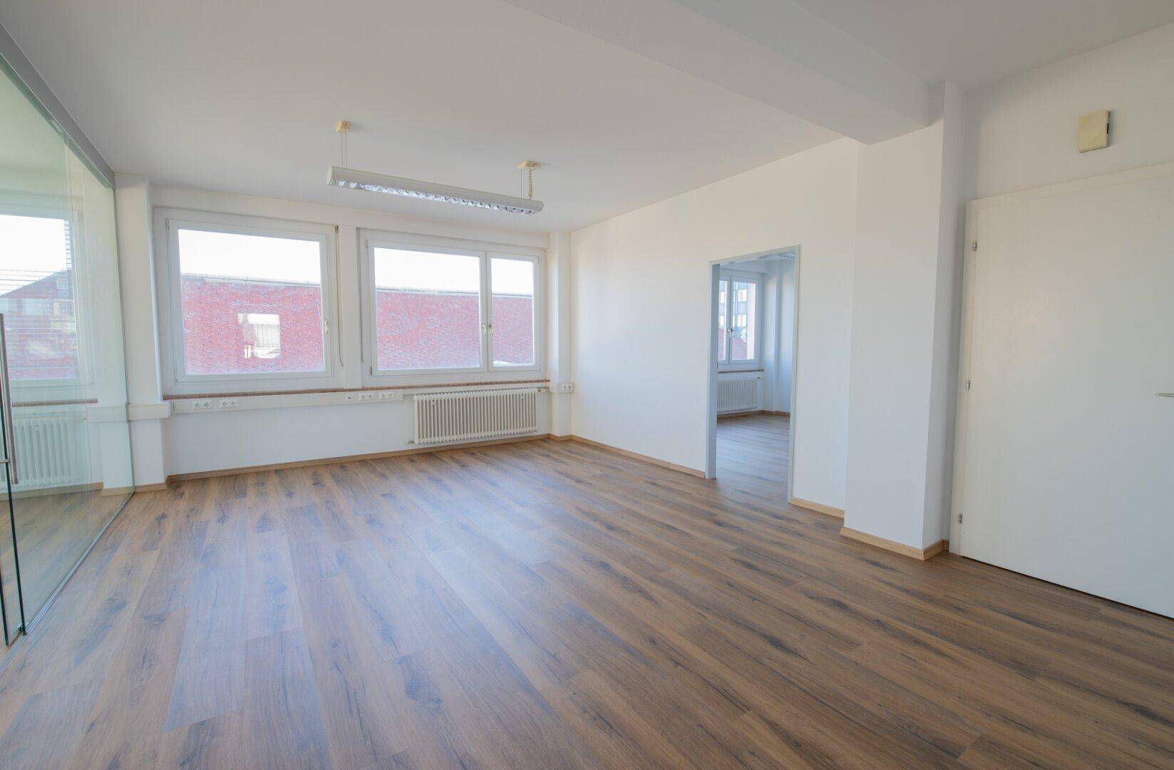 Leerer Büroraum mit hellen Wänden, Laminatboden und Blick auf die umliegenden Gebäude durch die Fenster.