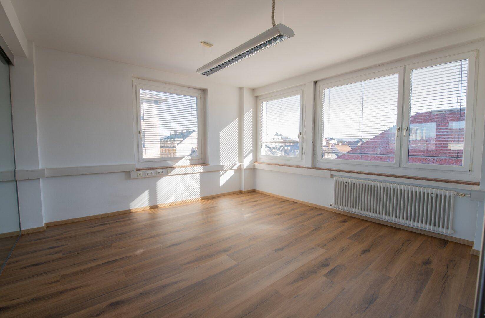 Sonniger Büroraum mit großen Fenstern, Jalousien und einem Heizkörper unter der Fensterbank.