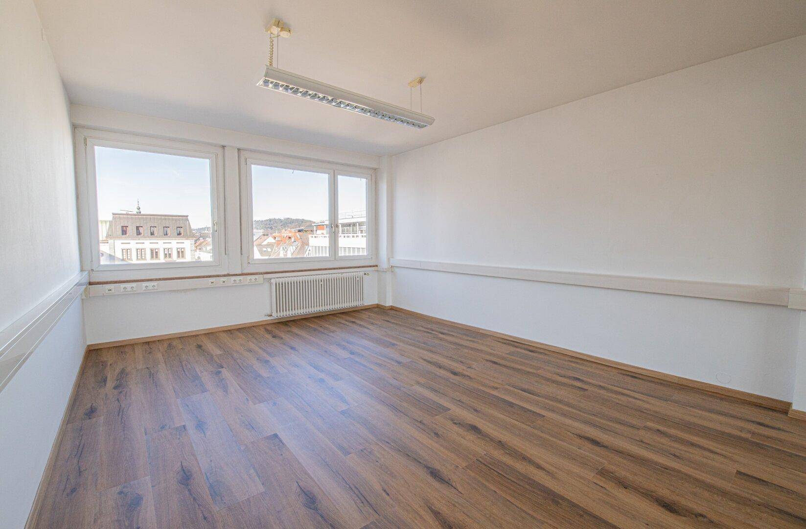 Leerer Büroraum mit Laminatboden, weißen Wänden und großen Fenstern, die viel Tageslicht hereinlassen.