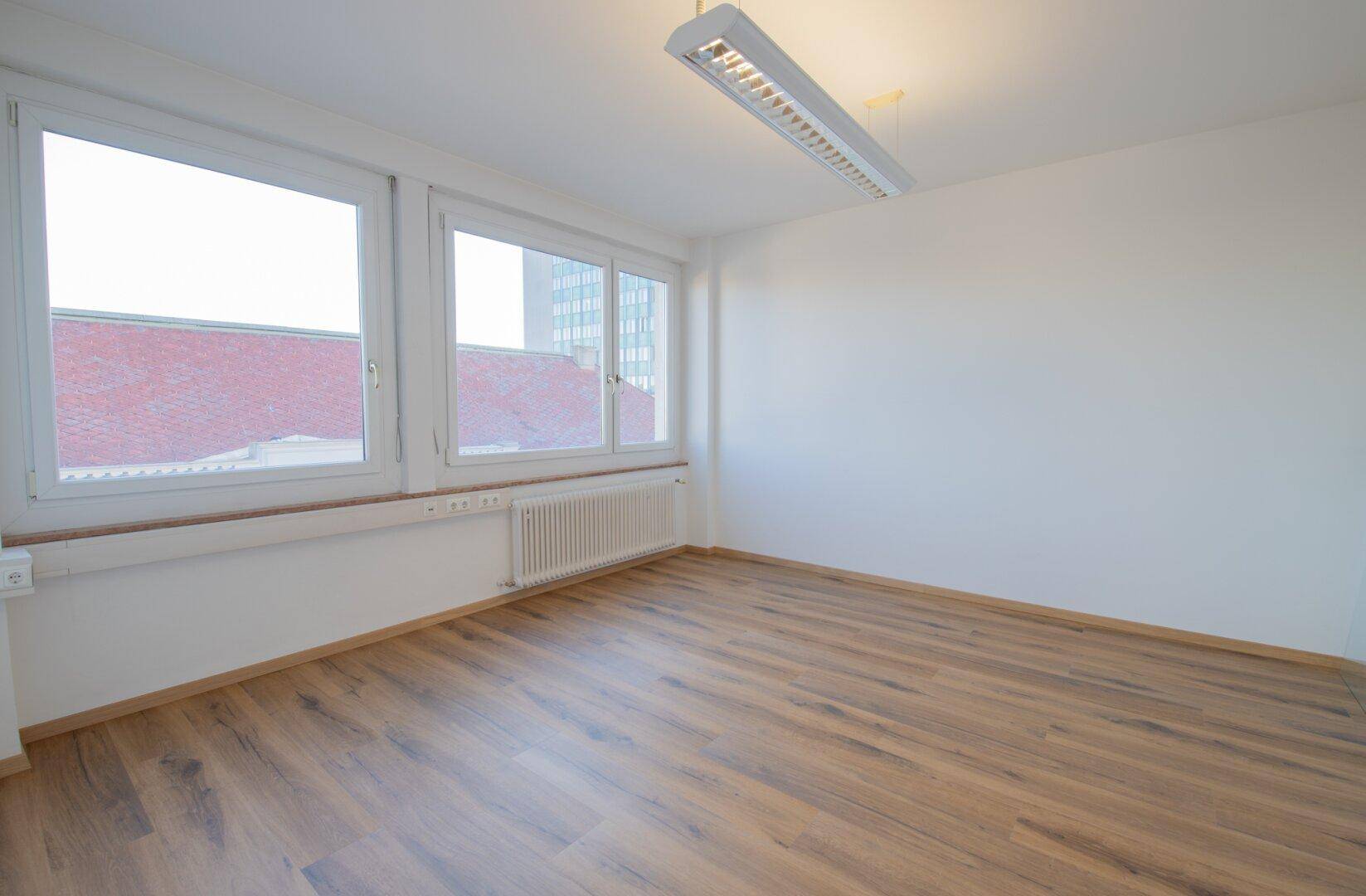 Heller Büroraum mit Laminatboden und großen Fenstern, die einen Blick auf die umliegenden Dächer bieten.