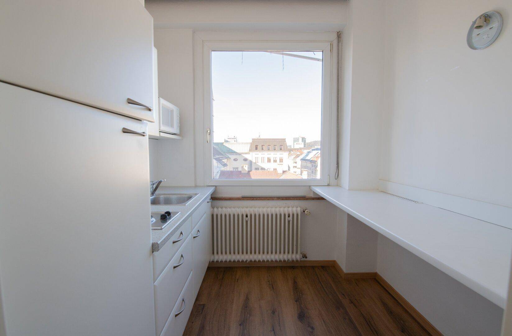 Kleine Küchenzeile mit weißen Schränken, Spüle und einem Fenster mit Blick auf die Stadt.