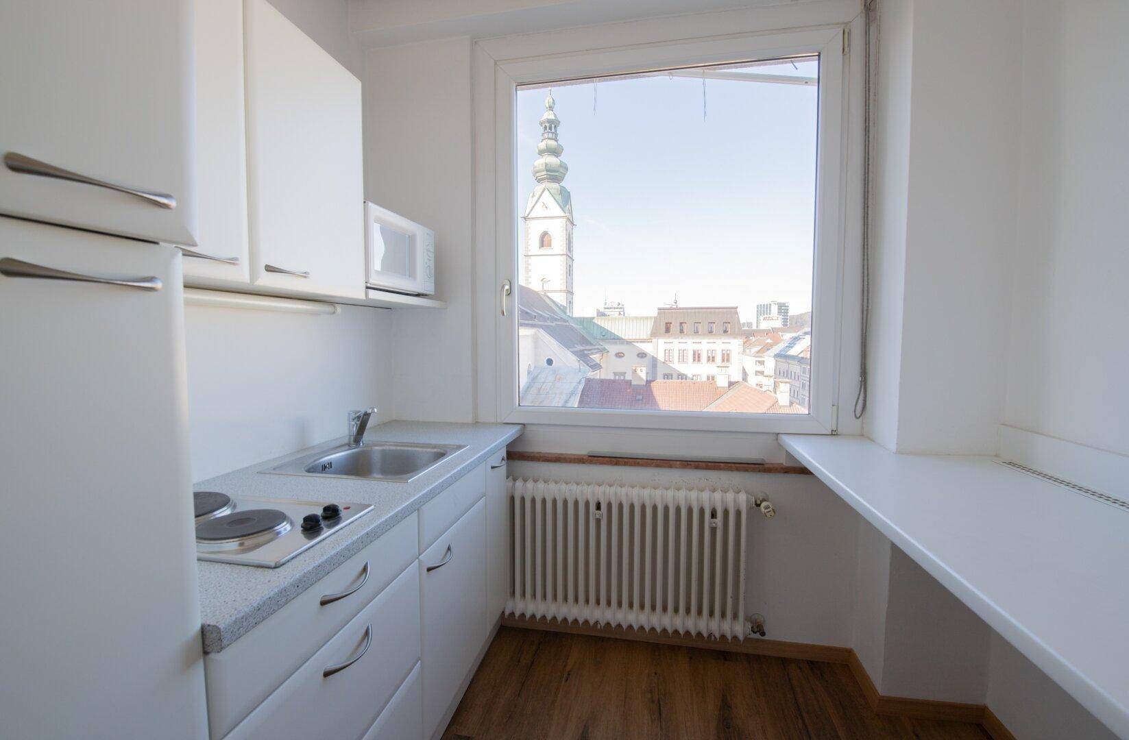 Kompakte Küchenzeile mit weißen Schränken, Kochfeld und einem Fenster mit Blick auf einen Kirchturm.