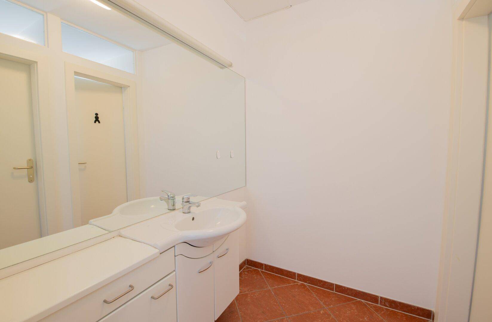 Badezimmer mit Waschbecken, großem Spiegel und Terrakotta-Fliesenboden in warmen Farbtönen.