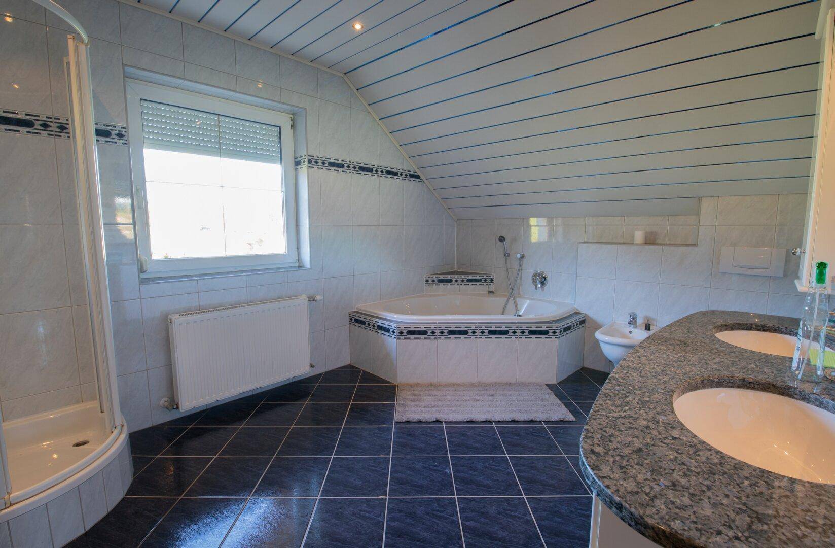 Gefliestes Badezimmer mit Eckbadewanne, Dusche, Doppelwaschbecken und Fenster im Dachgeschoss.
