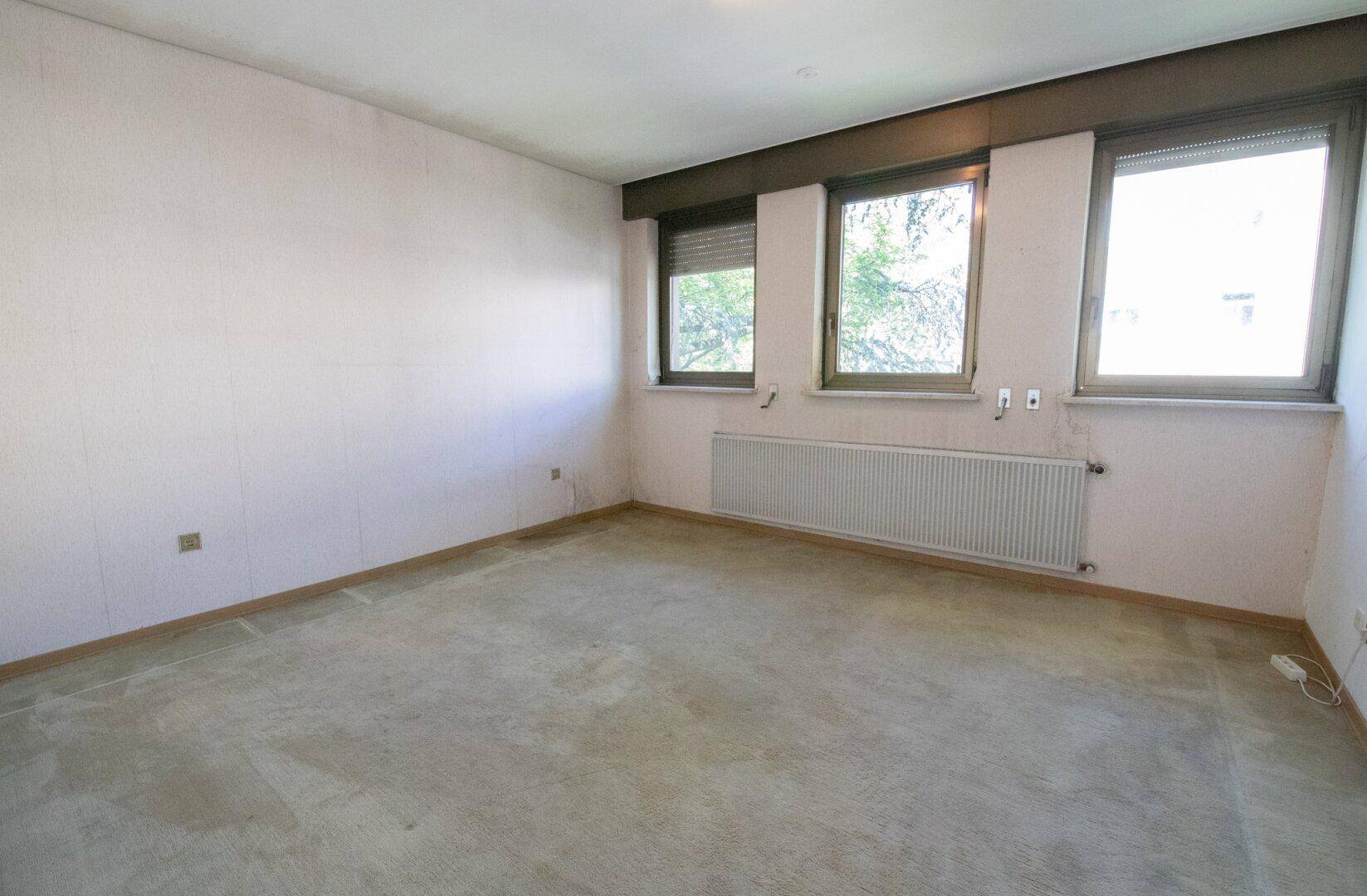 Schlafzimmer mit drei Fenstern, beigen Wänden und Teppichboden.