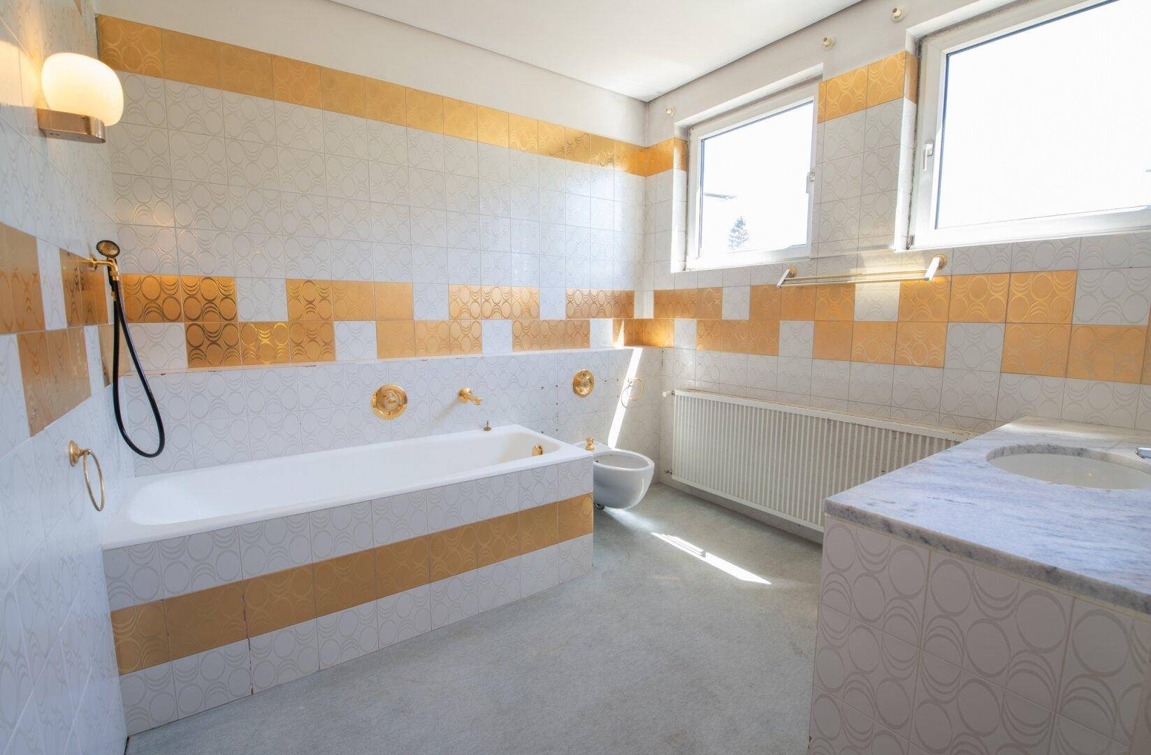 Elegantes Badezimmer mit gold-weißen Fliesen, Badewanne, Bidet und Doppelwaschbecken.