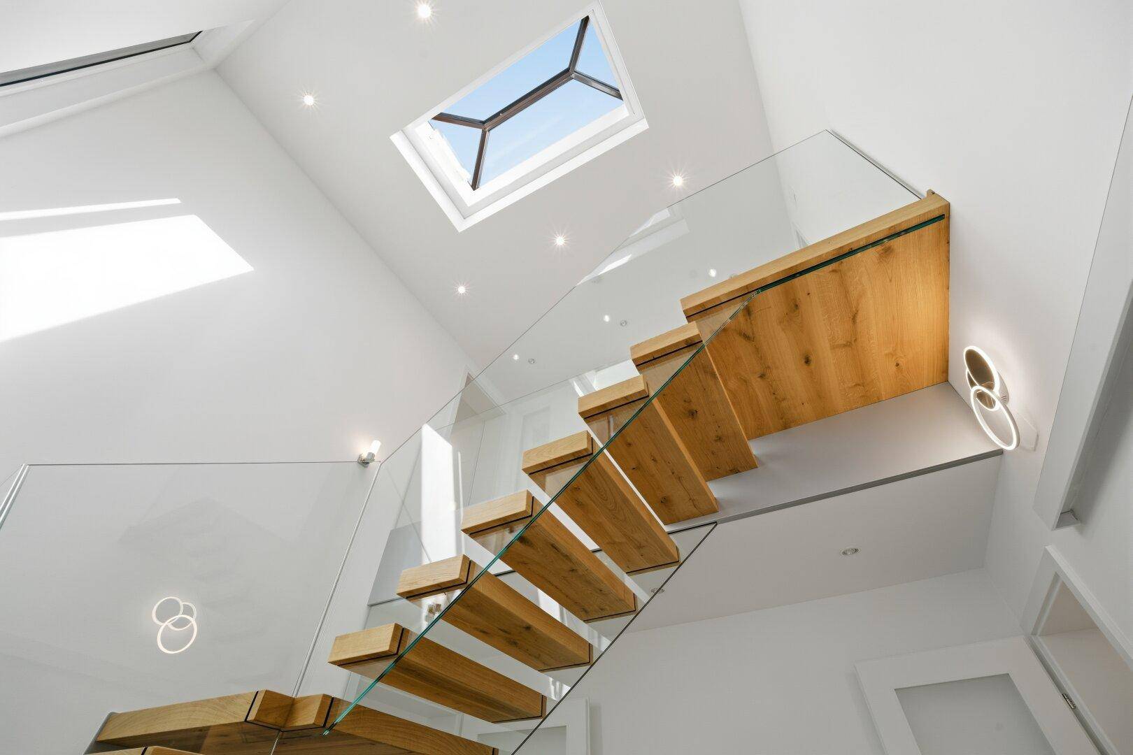 Helles Treppenhaus mit schwebenden Holzstufen, Glasgeländer und Oberlicht für viel Tageslicht.