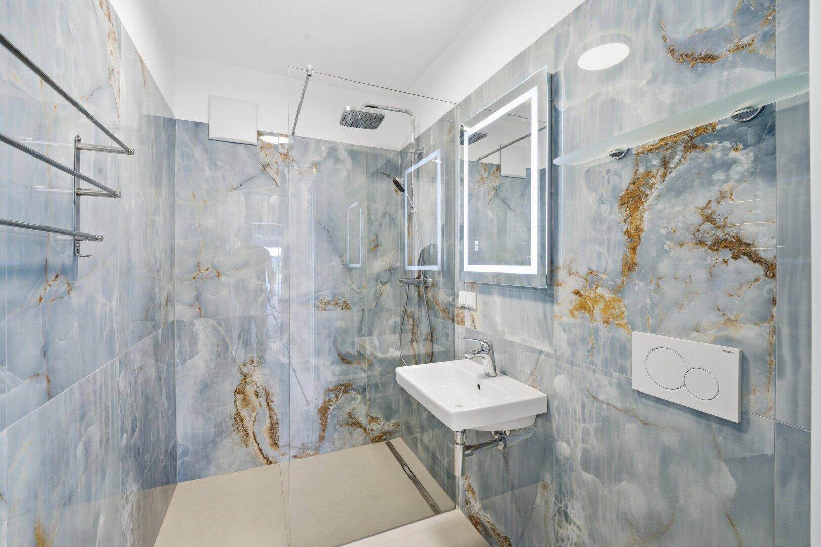 Modernes Badezimmer mit blauen Marmorfliesen, begehbarer Dusche, Einzelwaschbecken und beleuchtetem Spiegel.