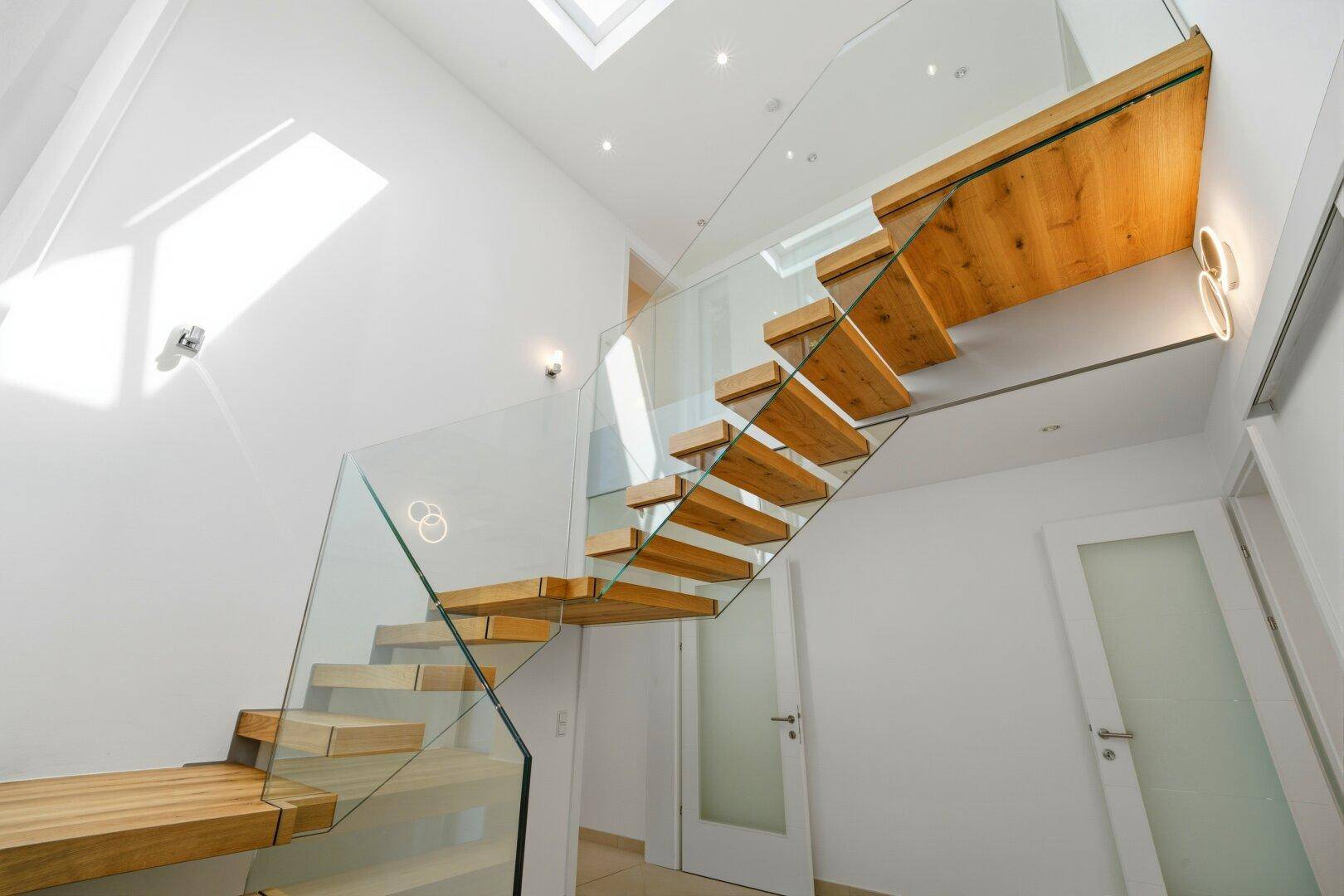 Elegantes Treppenhaus mit Holzstufen, Glasgeländer und Oberlicht, das den Raum mit Licht durchflutet.