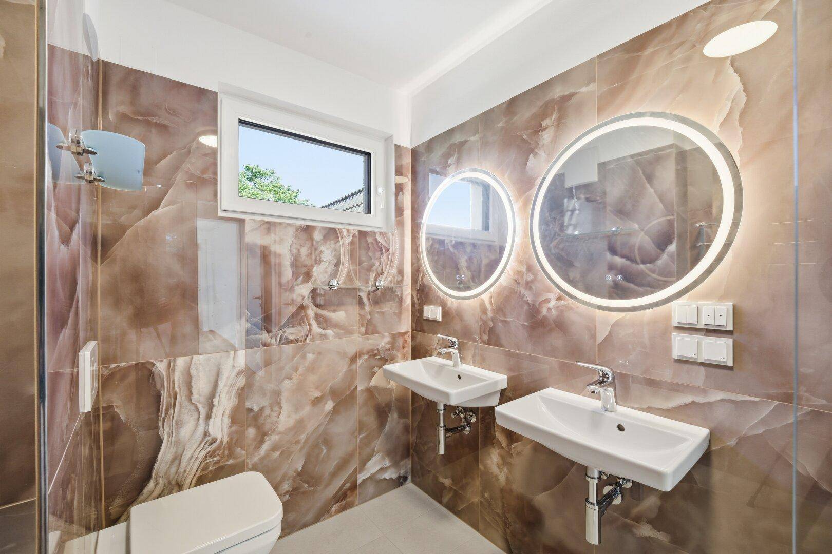 Stilvolles Badezimmer mit braunen Marmorfliesen, Doppelwaschbecken, WC und runden beleuchteten Spiegeln.