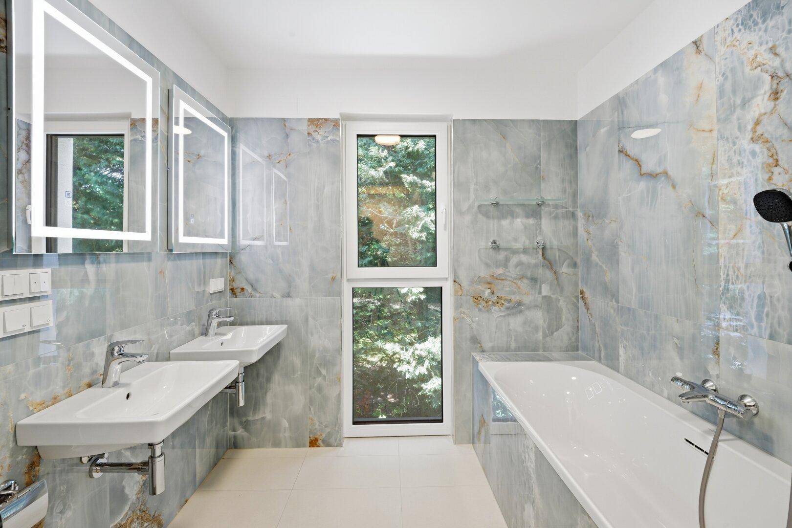 Stilvolles Badezimmer mit blauen Marmorfliesen, Doppelwaschbecken, Badewanne und Fenster mit Grünblick.