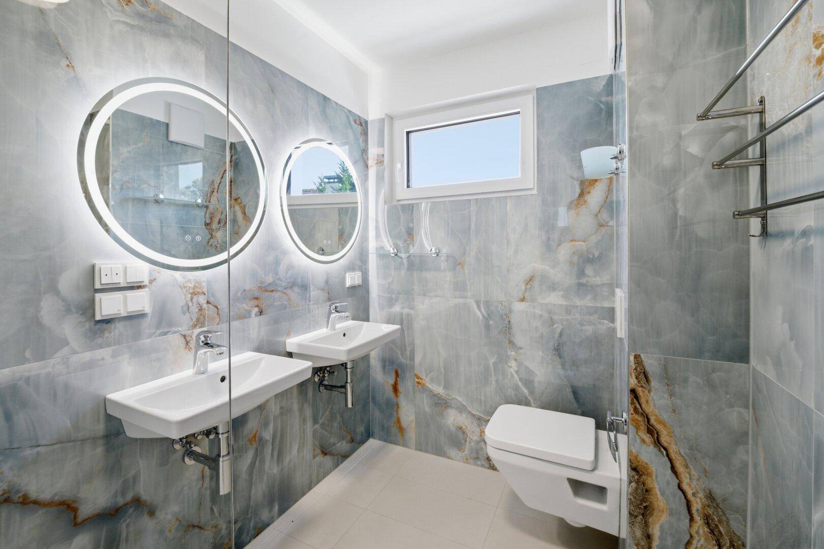 Elegantes Badezimmer mit blauen Marmorfliesen, Doppelwaschbecken, WC und runden beleuchteten Spiegeln.