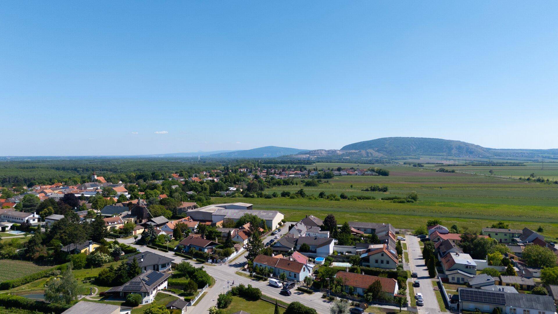 Weitläufige Luftaufnahme einer ländlichen Wohnsiedlung mit grünen Feldern und Hügeln im Hintergrund.