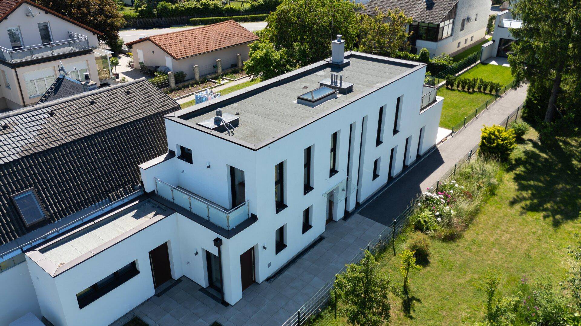 Moderne Villa mit Flachdach und großem Garten, umgeben von weiteren Wohnhäusern in grüner Umgebung.