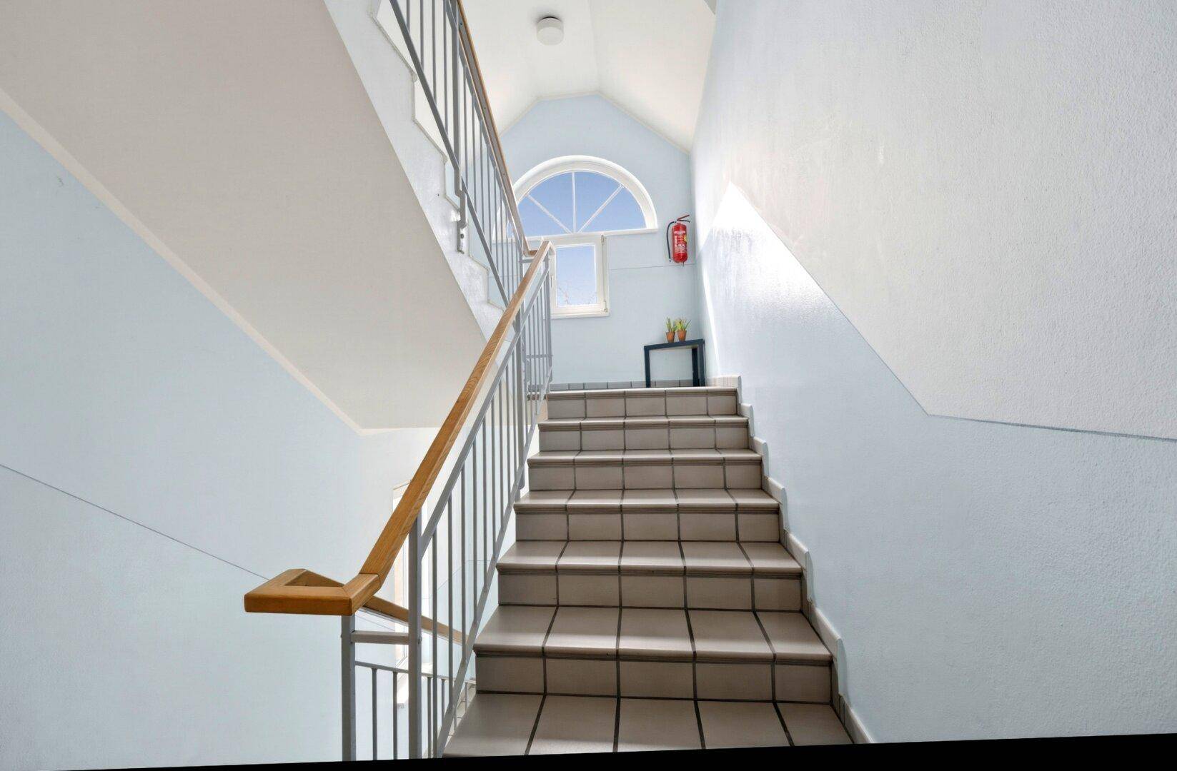Helles Treppenhaus mit gefliesten Stufen, Metallgeländer und einem Bogenfenster im oberen Bereich.