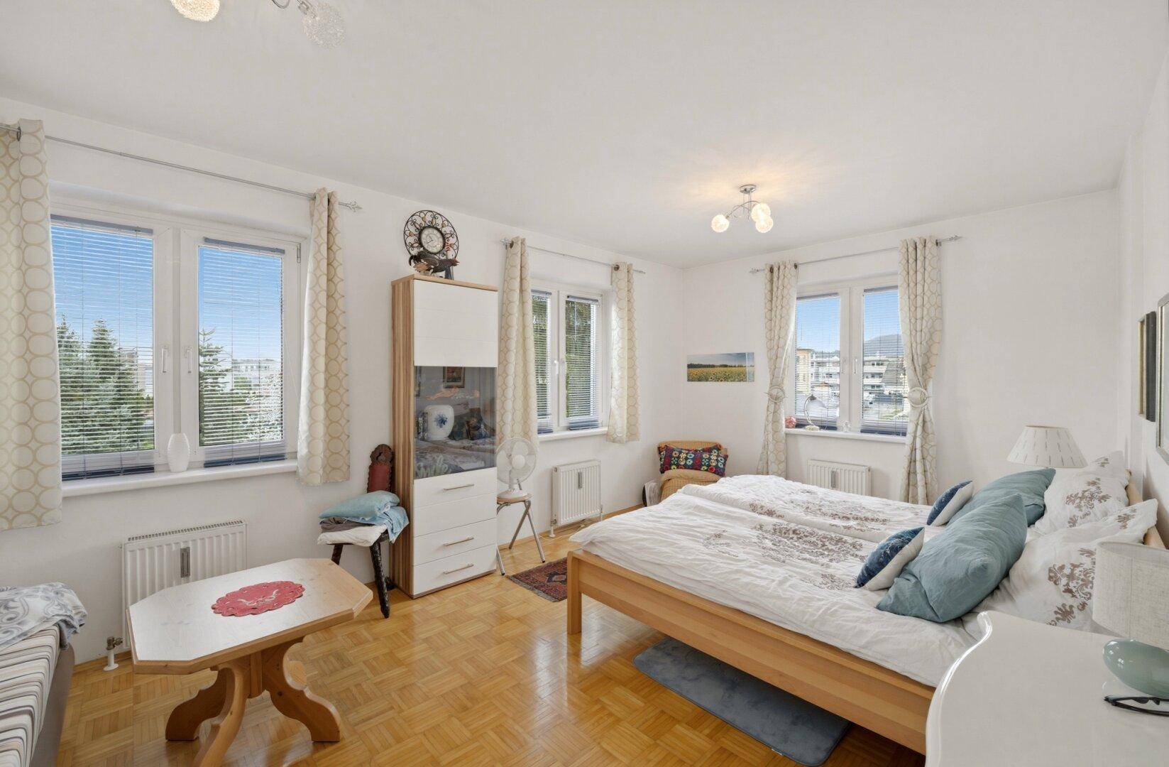 Helles Schlafzimmer mit Parkettboden, Doppelbett und mehreren Fenstern mit Blick ins Grüne.
