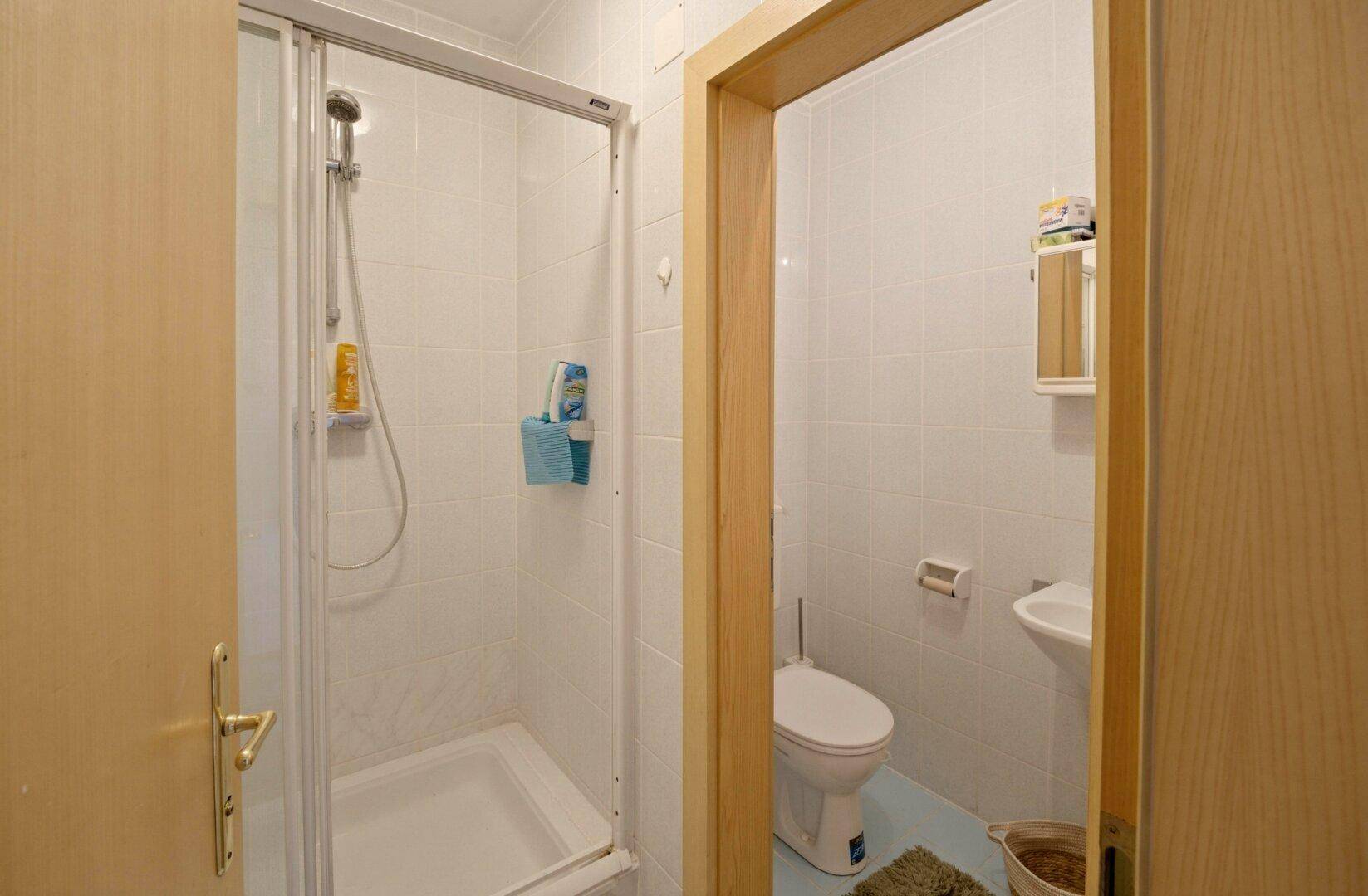 Kleines Badezimmer mit Duschkabine, WC und Waschbecken, komplett gefliest in Weiß.