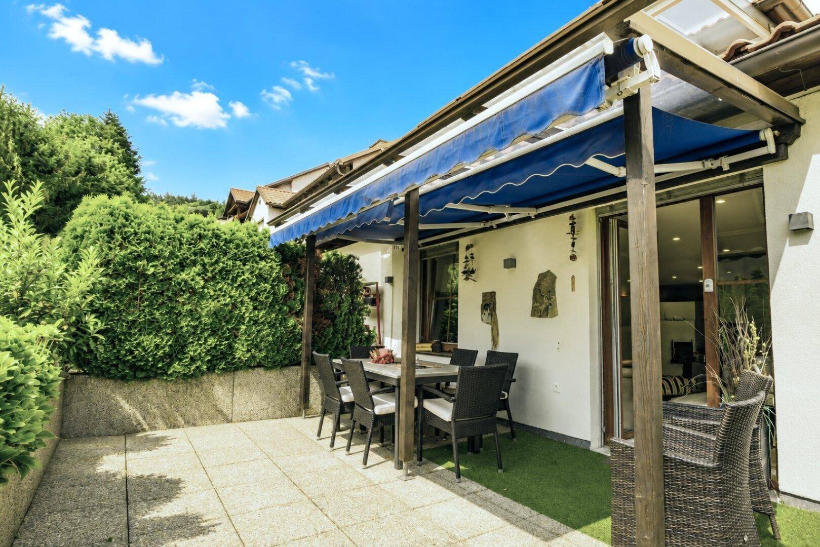 Geräumige Terrasse mit Essbereich und Markise, umgeben von gepflegten Hecken und Pflanzen.