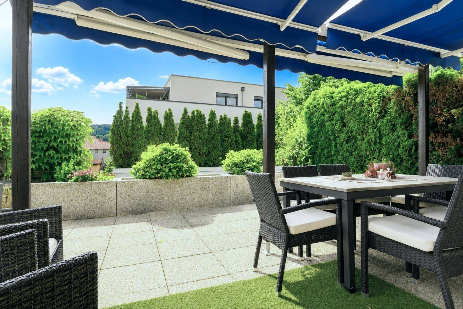 Große, sonnige Terrasse mit Esstisch und Lounge-Möbeln unter einer blauen Markise, umgeben von Grün.