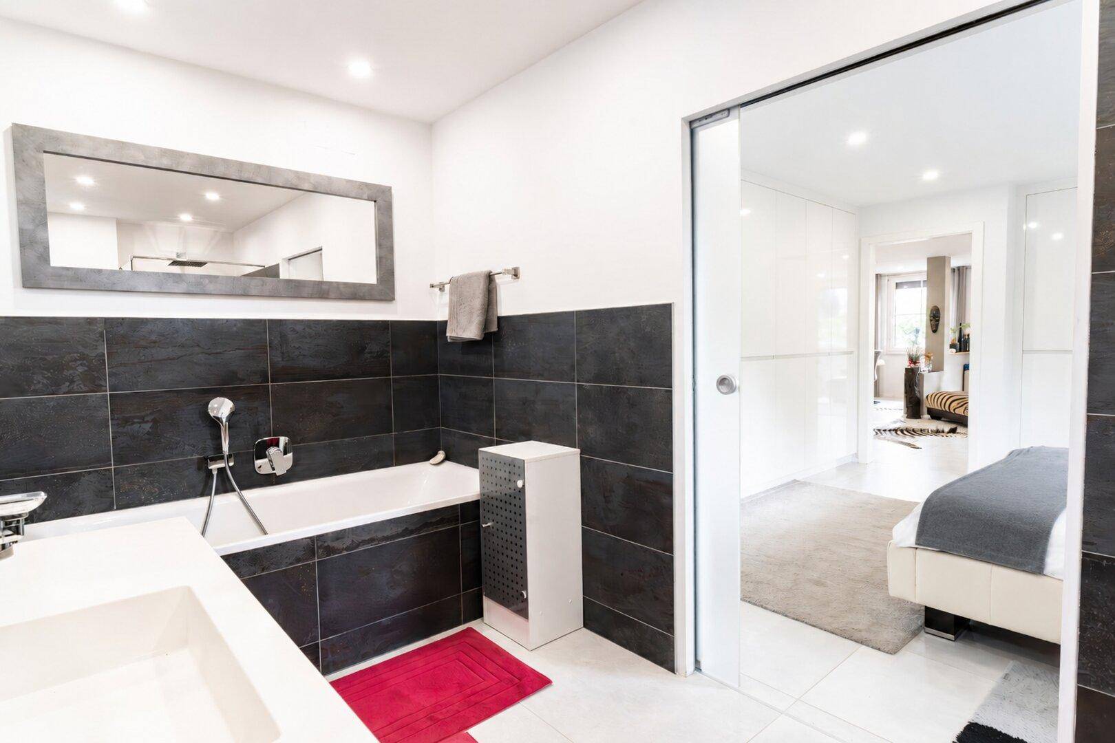 Modernes Badezimmer mit Badewanne, dunklen Fliesen und direktem Zugang zum Schlafzimmer.