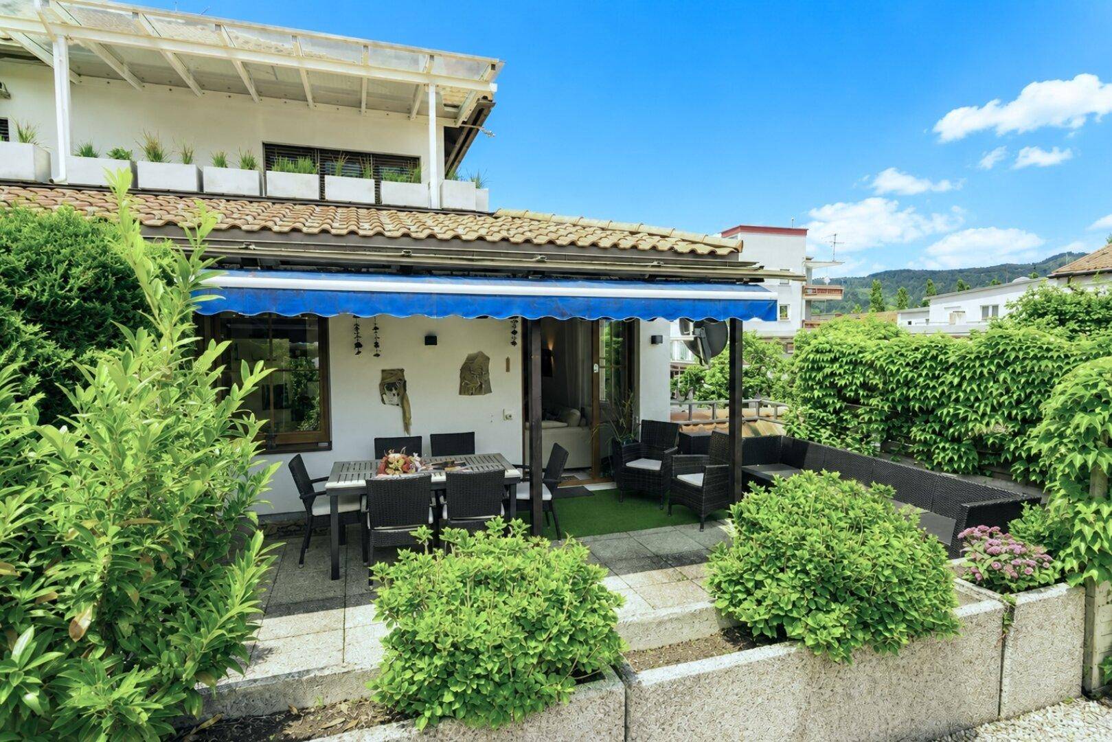 Sonnige Terrasse mit Esstisch, Lounge-Möbeln und blauer Markise, umgeben von grünen Pflanzen.