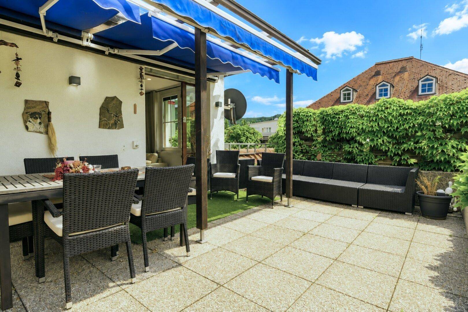 Weitläufige Terrasse mit Ess- und Lounge-Bereich, blauer Markise und Blick auf die Umgebung.