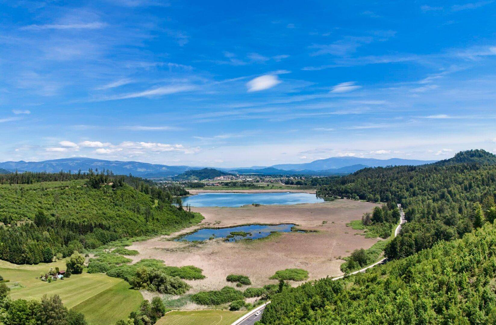 Weitläufige Landschaft mit einem See, umgeben von Wäldern und Bergen unter blauem Himmel.