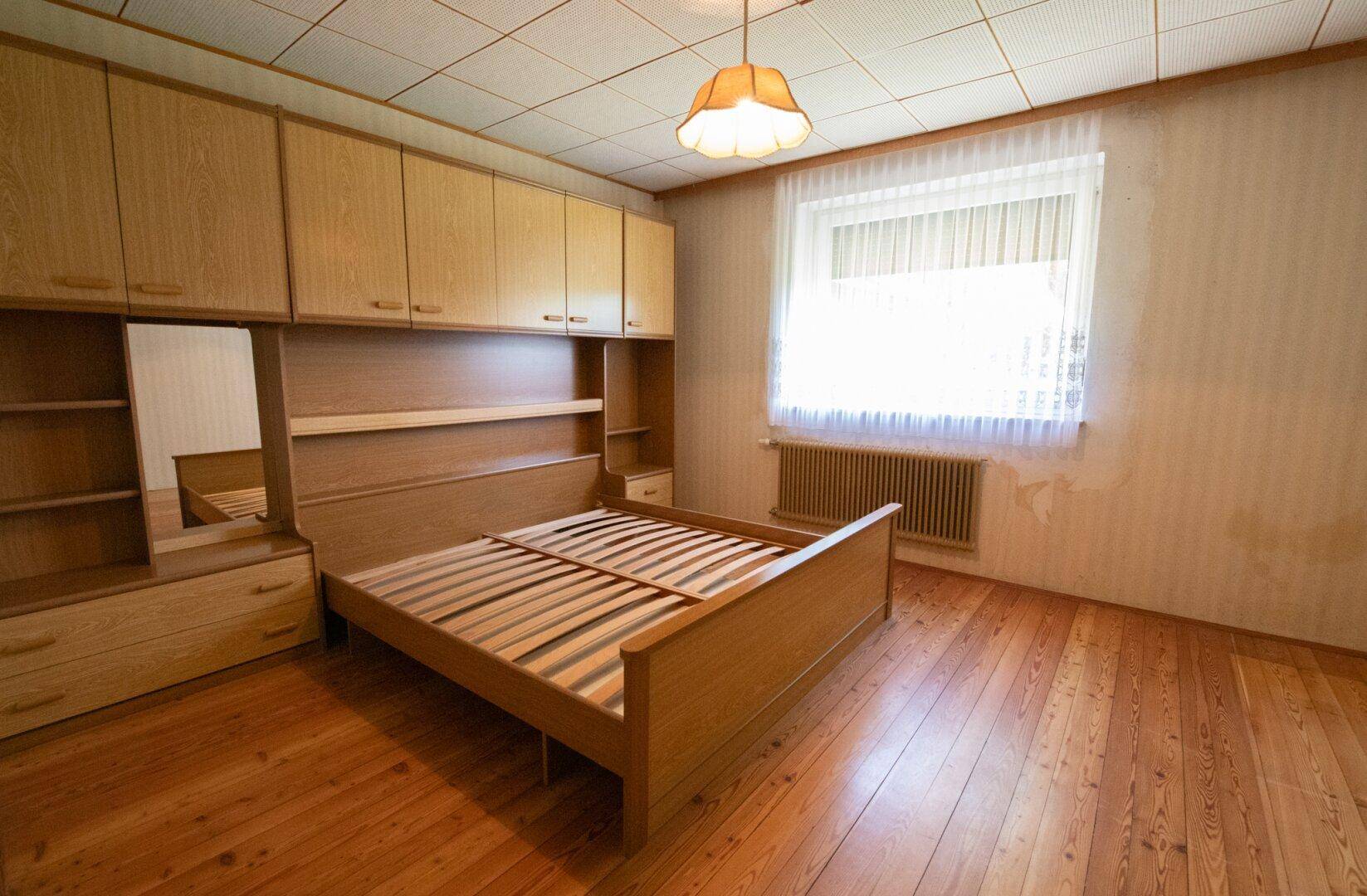 Schlafzimmer mit Holzmöbeln, Parkettboden und einem Fenster mit Blick ins Grüne.