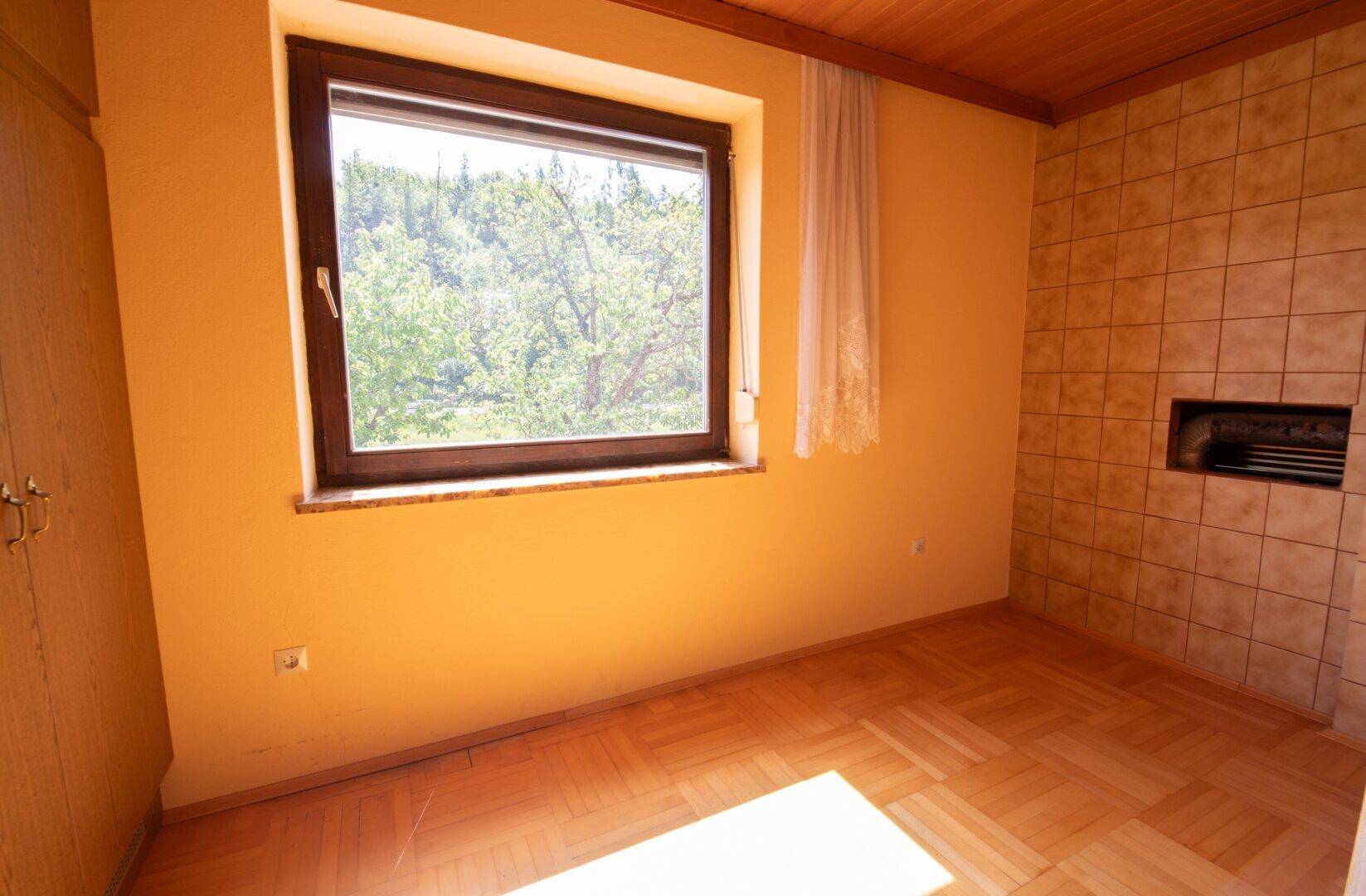 Heller Raum mit Parkettboden, großem Fenster und einer gefliesten Wand mit Kaminöffnung.