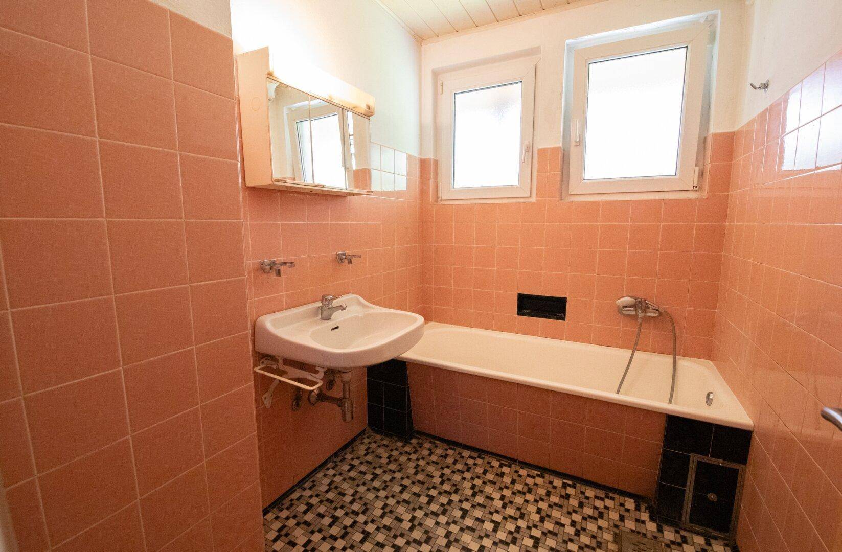 Badezimmer mit rosa Fliesen, Badewanne und zwei Fenstern für natürliche Belüftung.