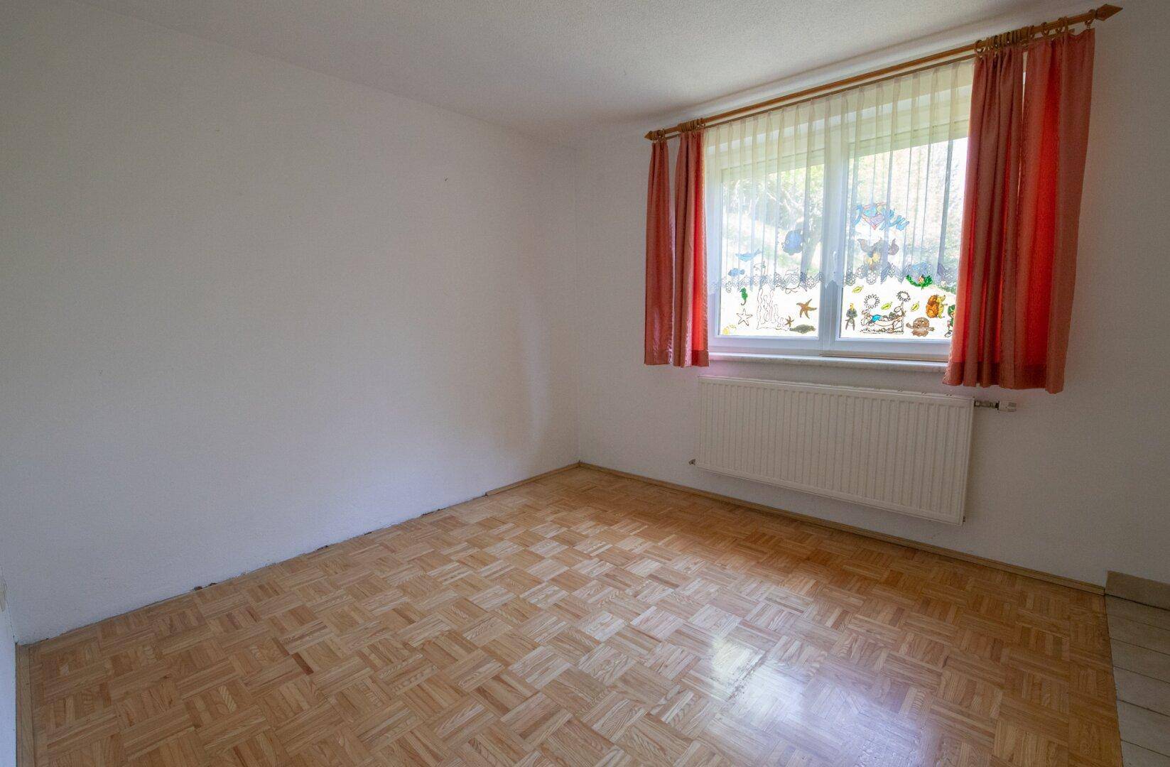 Helles Zimmer mit Parkettboden und großem Fenster, dekoriert mit bunten Vorhängen.