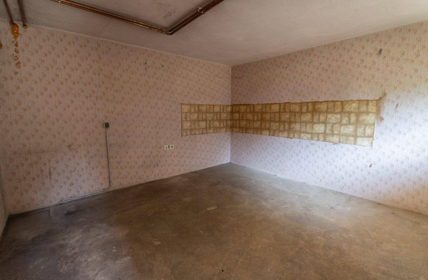 Unfertiger Kellerraum mit alter Tapete, Betonboden und sichtbaren Leitungen, ideal für Lagerzwecke.
