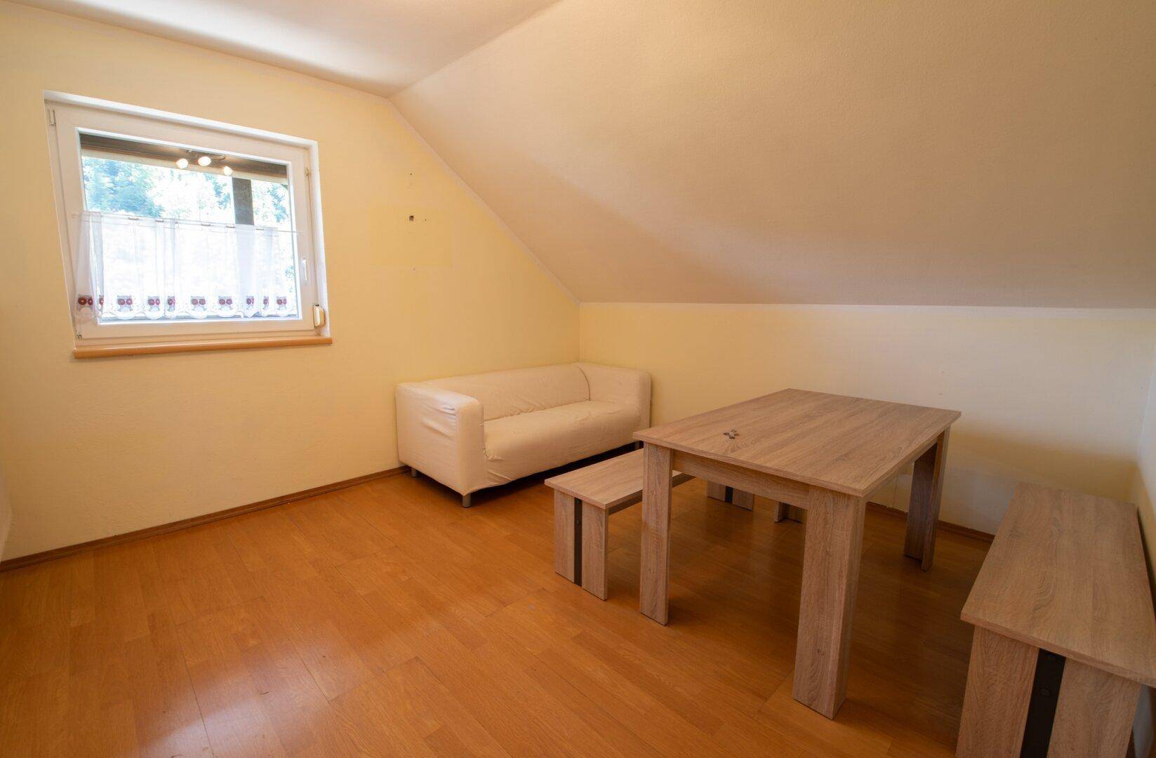Heller Dachgeschossraum mit Holzboden, Sofa und Esstisch unter einem Fenster.