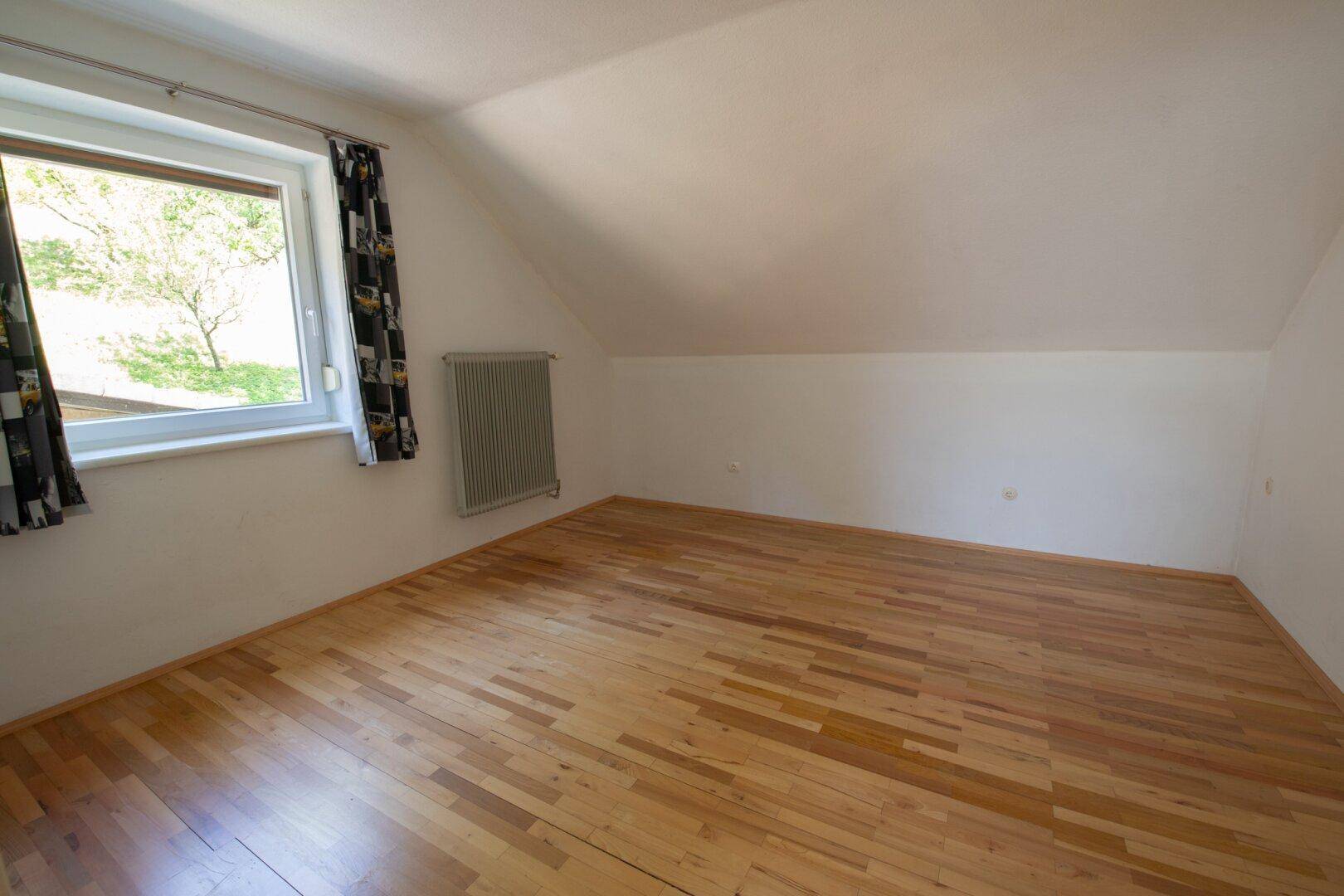 Helles Dachgeschosszimmer mit Holzboden und Fenster mit Blick ins Grüne.