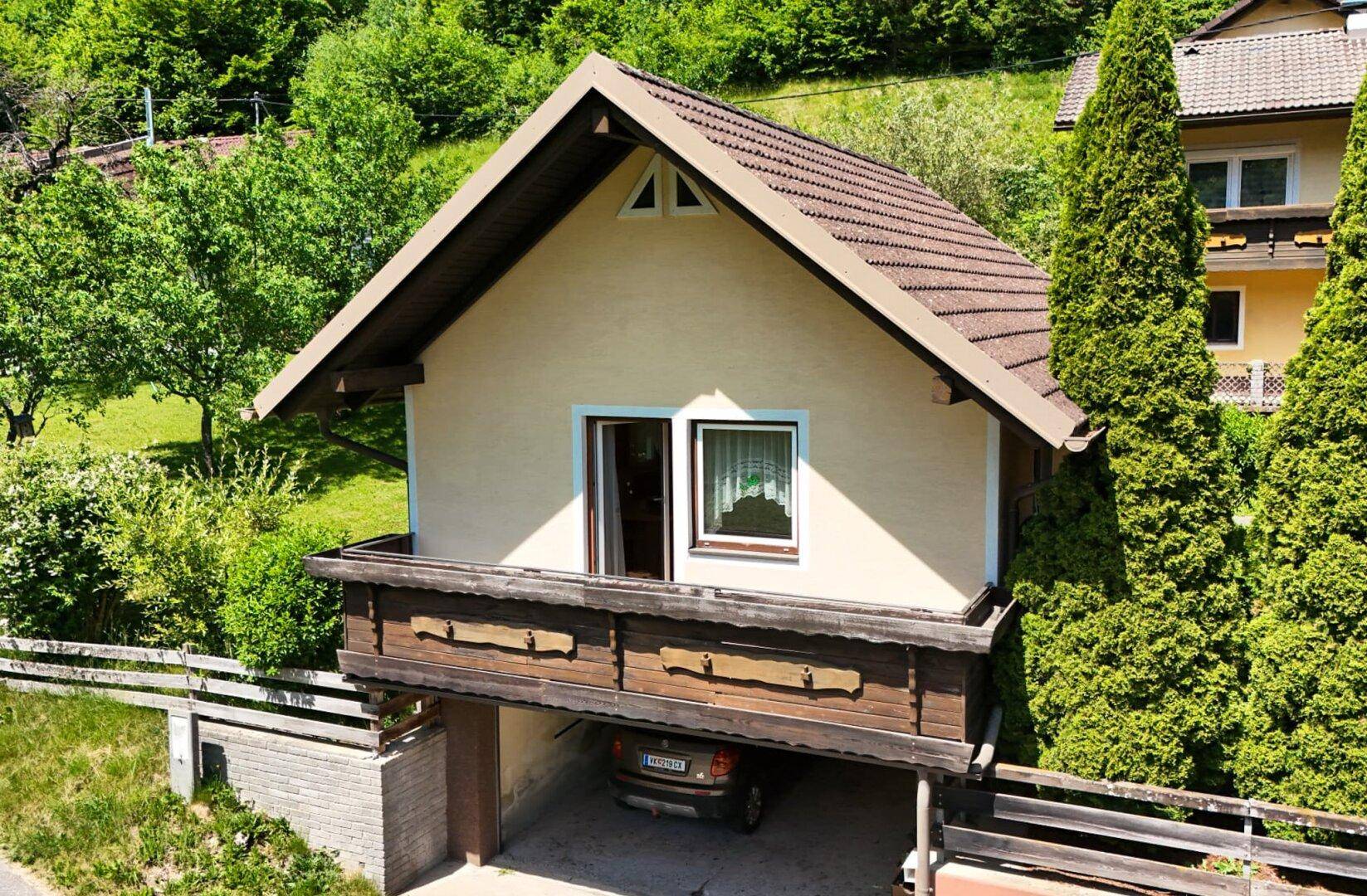 Kleines Haus mit traditionellem Holzbalkon und überdachtem Parkplatz in grüner Hanglage.