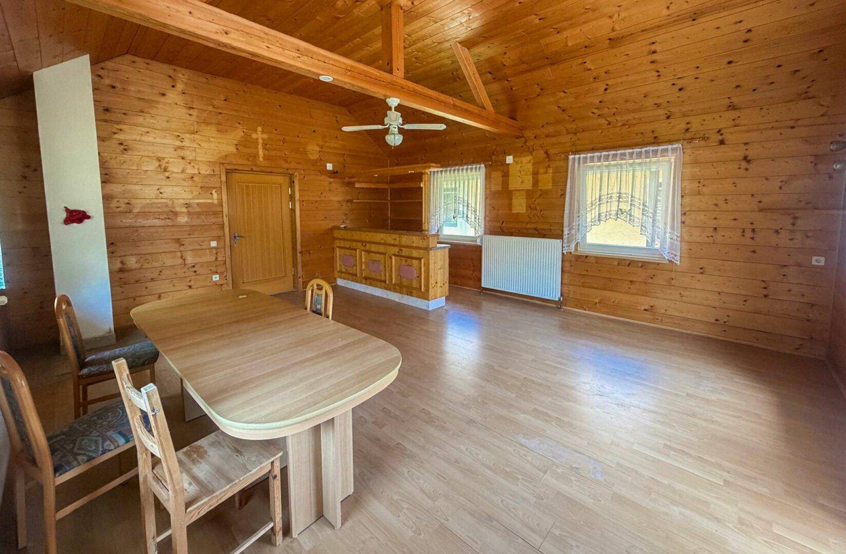 Rustikaler Wohnbereich mit Holzwänden, Esstisch und Deckenventilator in ländlicher Umgebung.