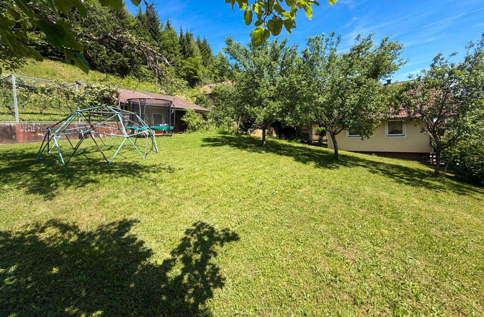 Großer, sonniger Garten mit Rasenfläche, Obstbäumen und Kinderspielgeräten in grüner Umgebung.