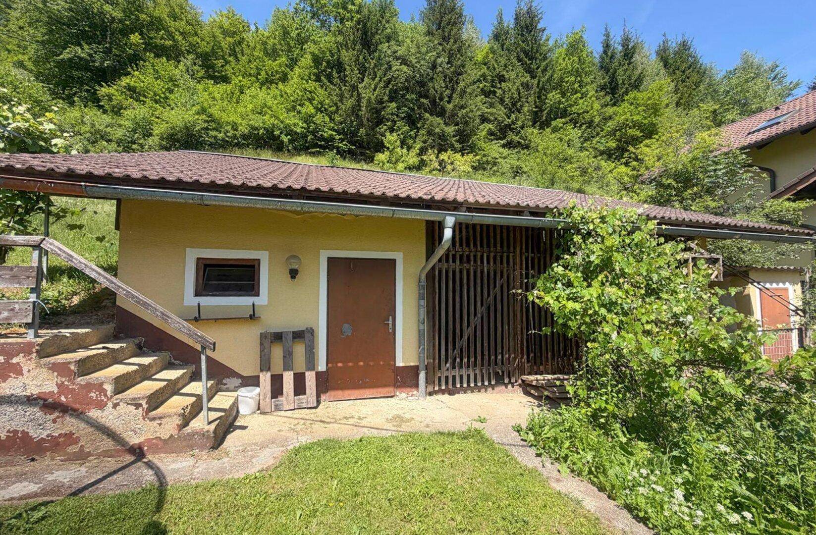 Kleines Nebengebäude im Garten mit Steintreppe und einfacher Fassade, umgeben von Grün.