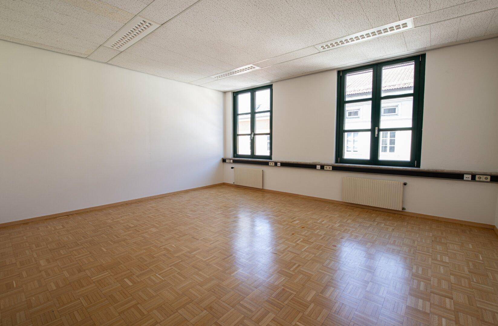 Helles Büro mit Fischgrätparkett und zwei großen Fenstern, die einen Blick nach draußen bieten.