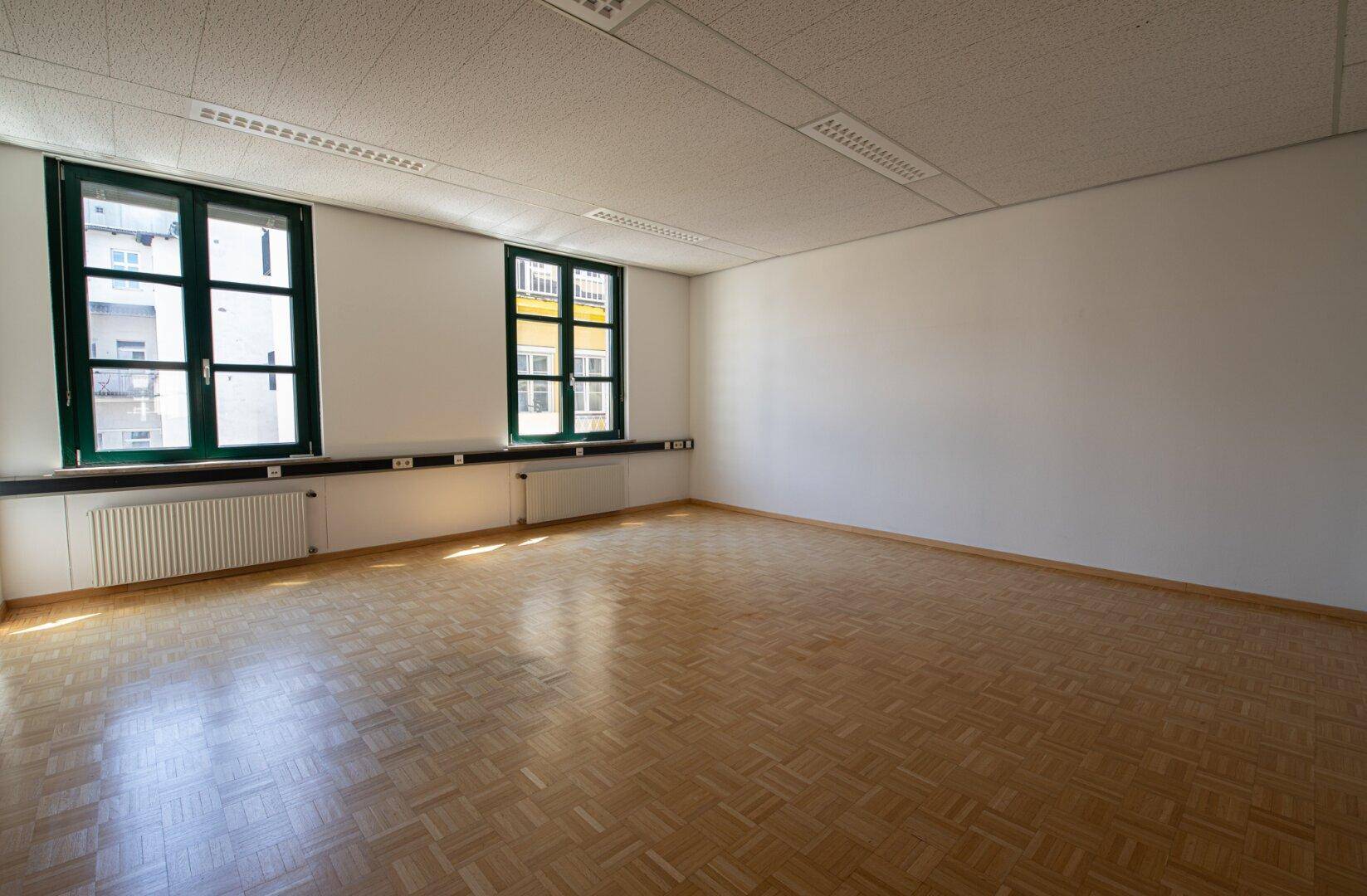 Großes, leeres Büro mit glänzendem Parkettboden und zwei Fenstern, die den Raum mit Licht füllen.