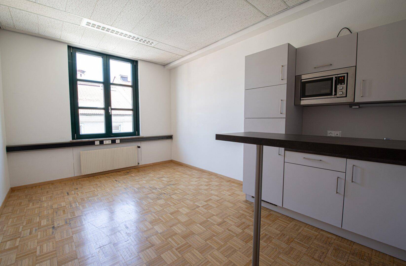 Kleiner Küchenbereich mit grauen Schränken, Mikrowelle und Parkettboden in einem hellen Raum.